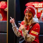 FootballR - NFL - Eine Frau lächelt und zeigt mit beiden Händen in die Kamera. Sie trägt ein rotes Kansas City Chiefs-Trikot und eine dazu passende Strickmütze. Hinter ihr im ChampionsHaus in Frankfurt hängen zwei Footballtrikots. Auf dem linken Trikot steht „MAHOMES“ und auf dem rechten „KELCE“ mit den jeweiligen Nummern. Diese Beschreibung wurde automatisch generiert.