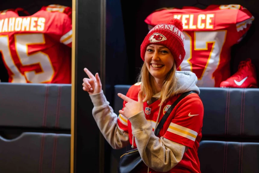 FootballR - NFL - Eine Frau lächelt und zeigt mit beiden Händen in die Kamera. Sie trägt ein rotes Kansas City Chiefs-Trikot und eine dazu passende Strickmütze. Hinter ihr im ChampionsHaus in Frankfurt hängen zwei Footballtrikots. Auf dem linken Trikot steht „MAHOMES“ und auf dem rechten „KELCE“ mit den jeweiligen Nummern. Diese Beschreibung wurde automatisch generiert.