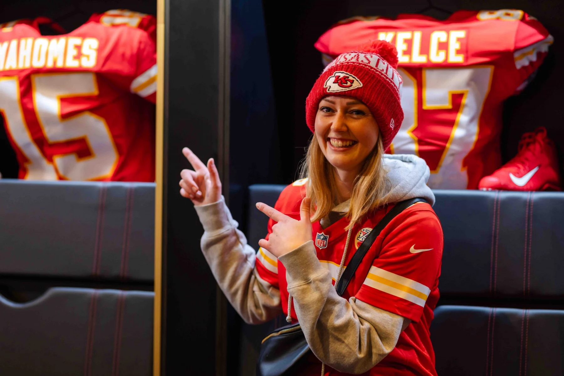 FootballR - NFL - Eine Frau lächelt und zeigt mit beiden Händen in die Kamera. Sie trägt ein rotes Kansas City Chiefs-Trikot und eine dazu passende Strickmütze. Hinter ihr im ChampionsHaus in Frankfurt hängen zwei Footballtrikots. Auf dem linken Trikot steht „MAHOMES“ und auf dem rechten „KELCE“ mit den jeweiligen Nummern. Diese Beschreibung wurde automatisch generiert.