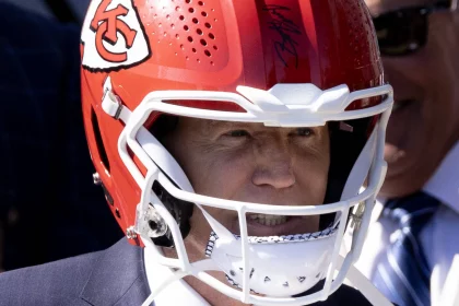 FootballR - NFL - US Präsident Joe Biden trägt einen roten Footballhelm mit dem Logo der Kansas City Chiefs an der Seite. Der Gesichtsschutz ist weiß und der Helm ist signiert. Die Person, die Joe Biden verblüffend ähnlich sieht, trägt ebenfalls einen dunklen Anzug, während von einer anderen Person im Hintergrund nur ein Teil zu sehen ist. Diese Beschreibung wurde automatisch generiert.