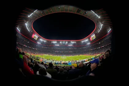 FootballR - NFL - Ein voll besetztes Footballstadion ist unter dem Abendhimmel beleuchtet. Das Spielfeld ist bereit für ein Spiel, Spieler und Offizielle sind versammelt. Fans füllen die Tribünen, viele halten Tickets für die NFL München 2024 in der Hand, während sie dem Ereignis entgegenfiebern. Der runde Rand des Stadions ist mit roten Lichtern hervorgehoben, was die elektrisierende Atmosphäre noch verstärkt. Diese Beschreibung wurde automatisch generiert.