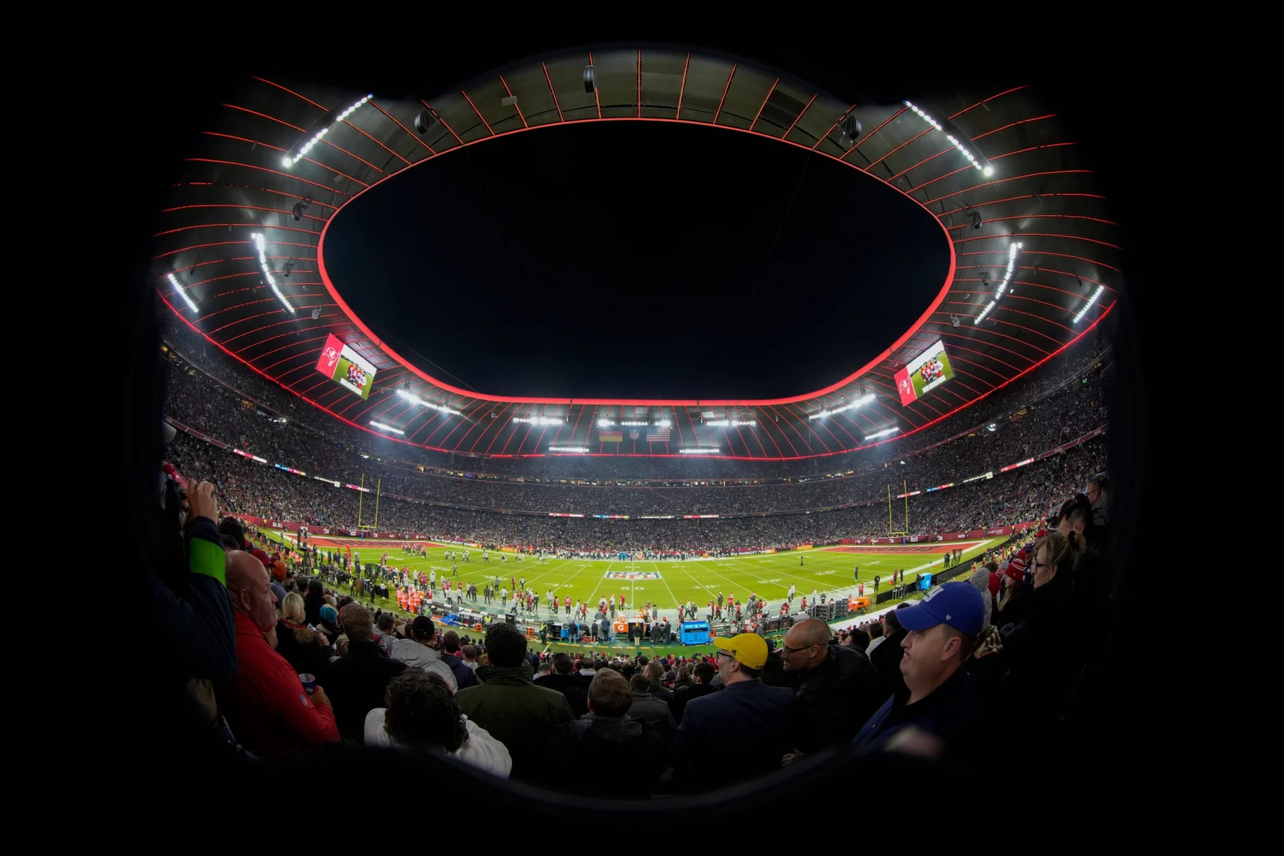 FootballR - NFL - Ein voll besetztes Footballstadion ist unter dem Abendhimmel beleuchtet. Das Spielfeld ist bereit für ein Spiel, Spieler und Offizielle sind versammelt. Fans füllen die Tribünen, viele halten Tickets für die NFL München 2024 in der Hand, während sie dem Ereignis entgegenfiebern. Der runde Rand des Stadions ist mit roten Lichtern hervorgehoben, was die elektrisierende Atmosphäre noch verstärkt. Diese Beschreibung wurde automatisch generiert.