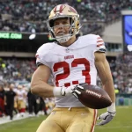 FootballR - NFL Madden NFL 25 - Christian McCaffrey - Ein Footballspieler in einem weißen Trikot mit roten und goldenen Akzenten, der Nummer 23 und einem goldenen Helm sprintet mit einem Football in der Hand über das Spielfeld. Im Hintergrund ist eine verschwommene Zuschauermenge im Stadion zu sehen, in der Ferne sind einige grüne Schilder zu sehen, die an das Cover von Madden NFL 25 erinnern. Diese Beschreibung wurde automatisch generiert.