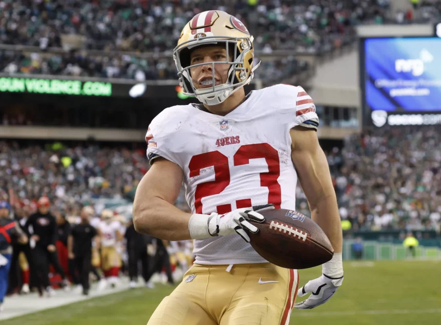 FootballR - NFL Madden NFL 25 - Christian McCaffrey - Ein Footballspieler in einem weißen Trikot mit roten und goldenen Akzenten, der Nummer 23 und einem goldenen Helm sprintet mit einem Football in der Hand über das Spielfeld. Im Hintergrund ist eine verschwommene Zuschauermenge im Stadion zu sehen, in der Ferne sind einige grüne Schilder zu sehen, die an das Cover von Madden NFL 25 erinnern. Diese Beschreibung wurde automatisch generiert.