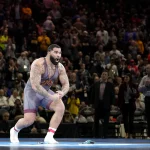 FootballR - NFL - Buffalo Bills - Ein muskulöser Wrestler in einem grauen Unterhemd mit kastanienbraunen und goldenen Akzenten feiert energisch auf der Matte und erinnert an Gable Steveson. Seine zahlreichen Tattoos auf seinen Armen und seiner oberen Brust fallen auf. Im Hintergrund zeigt eine große, jubelnde Menge in heller Kleidung eine Mischung aus konzentrierten und aufgeregten Ausdrücken. Diese Beschreibung wurde automatisch generiert.