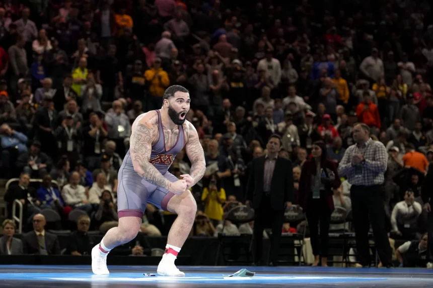 FootballR - NFL - Buffalo Bills - Ein muskulöser Wrestler in einem grauen Unterhemd mit kastanienbraunen und goldenen Akzenten feiert energisch auf der Matte und erinnert an Gable Steveson. Seine zahlreichen Tattoos auf seinen Armen und seiner oberen Brust fallen auf. Im Hintergrund zeigt eine große, jubelnde Menge in heller Kleidung eine Mischung aus konzentrierten und aufgeregten Ausdrücken. Diese Beschreibung wurde automatisch generiert.