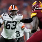 FootballR - NFL - Ein Spieler der Cleveland Browns in weißem Trikot und orangefarbenem Helm (Nummer 63) versucht, Lonnie Phelps von den Washington Commanders in roter Uniform (Nummer 78) während eines NFL-Spiels zu blockieren. Die Szene fängt die intensive Action auf dem Spielfeld ein, mit verschwommenen Zuschauern im Hintergrund. Diese Beschreibung wurde automatisch generiert.