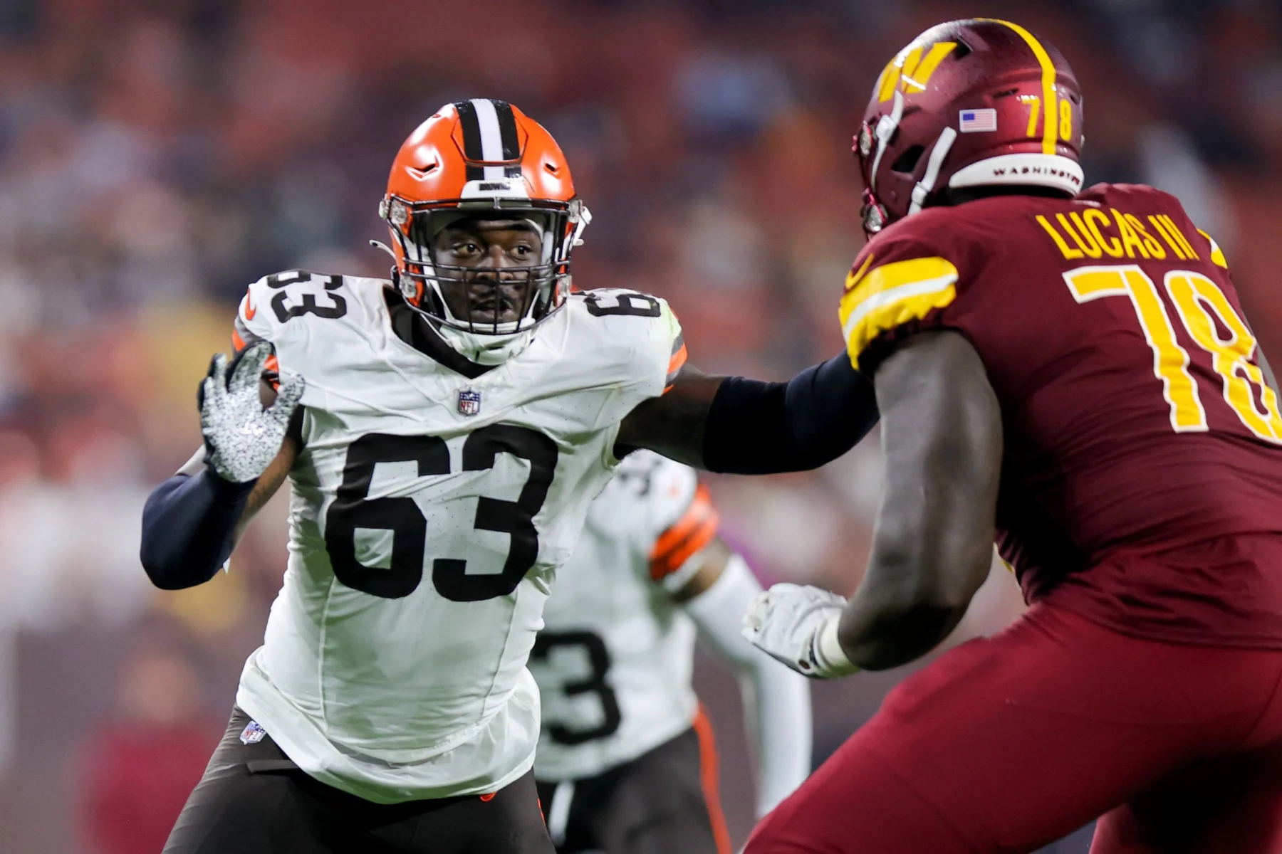 FootballR - NFL - Ein Spieler der Cleveland Browns in weißem Trikot und orangefarbenem Helm (Nummer 63) versucht, Lonnie Phelps von den Washington Commanders in roter Uniform (Nummer 78) während eines NFL-Spiels zu blockieren. Die Szene fängt die intensive Action auf dem Spielfeld ein, mit verschwommenen Zuschauern im Hintergrund. Diese Beschreibung wurde automatisch generiert.