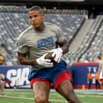 FootballR - NFL - Ein Footballspieler, der ein hellgraues Shirt mit der Aufschrift „GIANTS“ in blauen Buchstaben und rote Shorts trägt und an Darren Waller, den Tight End der New York Giants, erinnert, hält einen Football in der Hand und läuft über ein grünes Feld. Im Hintergrund ist ein Stadion mit Reihen leerer Sitze und ein paar Menschen in Sportkleidung in der Nähe der Spielfeldmarkierungen und Torpfosten zu sehen. Diese Beschreibung wurde automatisch generiert.
