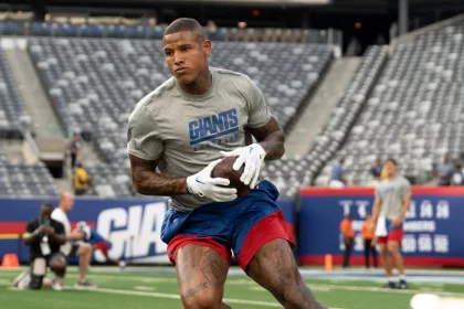 FootballR - NFL - Ein Footballspieler, der ein hellgraues Shirt mit der Aufschrift „GIANTS“ in blauen Buchstaben und rote Shorts trägt und an Darren Waller, den Tight End der New York Giants, erinnert, hält einen Football in der Hand und läuft über ein grünes Feld. Im Hintergrund ist ein Stadion mit Reihen leerer Sitze und ein paar Menschen in Sportkleidung in der Nähe der Spielfeldmarkierungen und Torpfosten zu sehen. Diese Beschreibung wurde automatisch generiert.