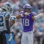 FootballR - NFL - Justin Jefferson - Ein Footballspieler in lila-weißer Uniform, Nummer 18, zeigt jubelnd nach vorne. Er wird von zwei Spielern in weiß-schwarzen Uniformen mit blauen Akzenten flankiert, einer von ihnen trägt die Nummer 42 auf seinem Trikot. Im Hintergrund sind eine verschwommene Menschenmenge und ein Stadion zu sehen, was auf eine Spielsituation hindeutet. Diese Beschreibung wurde automatisch generiert.