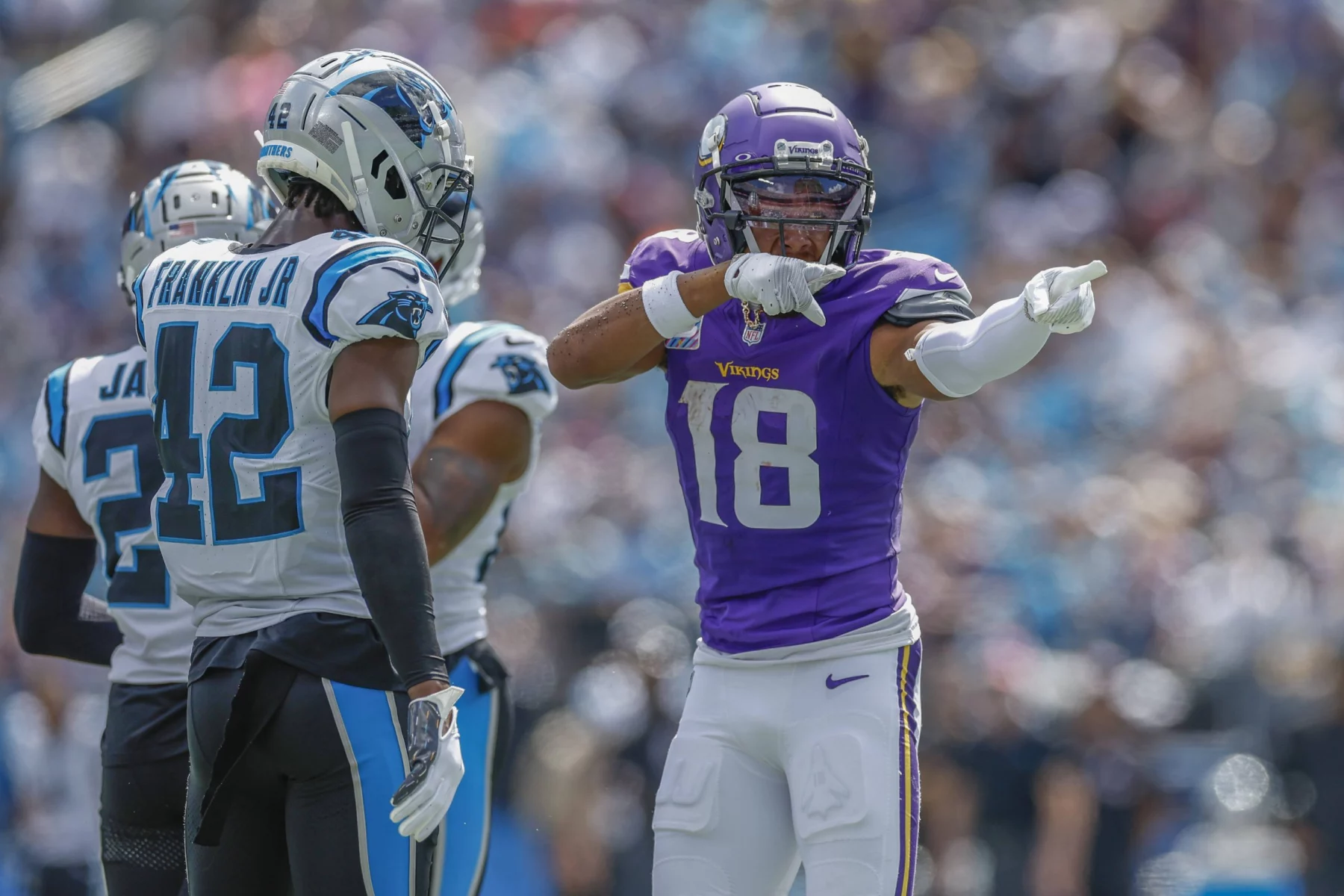 FootballR - NFL - Justin Jefferson - Ein Footballspieler in lila-weißer Uniform, Nummer 18, zeigt jubelnd nach vorne. Er wird von zwei Spielern in weiß-schwarzen Uniformen mit blauen Akzenten flankiert, einer von ihnen trägt die Nummer 42 auf seinem Trikot. Im Hintergrund sind eine verschwommene Menschenmenge und ein Stadion zu sehen, was auf eine Spielsituation hindeutet. Diese Beschreibung wurde automatisch generiert.