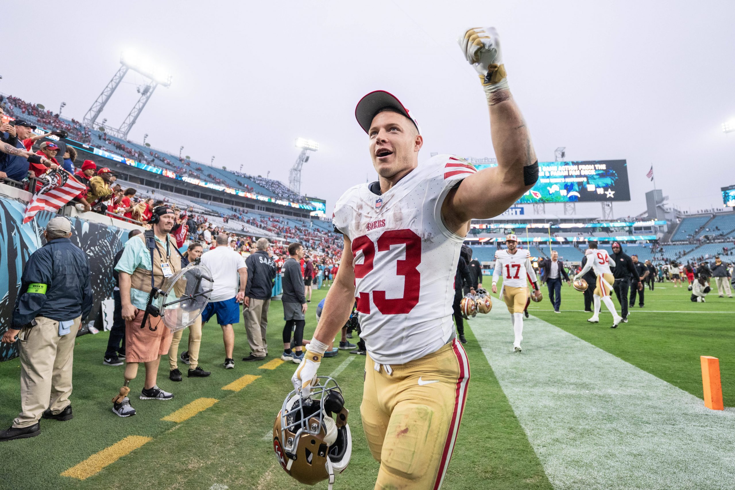 FootballR - NFL - Ein Footballspieler in einem weißen Trikot mit roten Nummern und goldenen Hosen verlässt das Feld, streckt triumphierend den Arm in die Höhe und hält seinen Helm in der Hand. Hinter ihm sind andere Spieler, Fotografen und Zuschauer zu sehen. Die Tribünen des Stadions sind voller jubelnder Fans, während auf großen Bildschirmen Christian McCaffrey, der Star der San Francisco 49ers, seinen jüngsten Sieg feiert, zu sehen ist. Diese Beschreibung wurde automatisch generiert.
