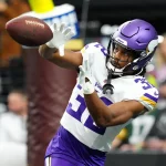 FootballR - NFL - Running Backs - Fantasy Football Geheimtipps 2024 Ty Chandler - Ein Footballspieler in der Uniform der Minnesota Vikings, einschließlich lila Helm und weißer Trikotnummer 32, fängt mit beiden Händen einen Football. Im Hintergrund ist ein verschwommenes Stadion mit Zuschauern zu sehen. In Aktion mit intensivem Fokus auf den Ball aufgenommen, verkörpert er einen der geheimsten Fantasy Football-Geheimtipps. Diese Beschreibung wurde automatisch generiert.