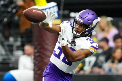 FootballR - NFL - Running Backs - Fantasy Football Geheimtipps 2024 Ty Chandler - Ein Footballspieler in der Uniform der Minnesota Vikings, einschließlich lila Helm und weißer Trikotnummer 32, fängt mit beiden Händen einen Football. Im Hintergrund ist ein verschwommenes Stadion mit Zuschauern zu sehen. In Aktion mit intensivem Fokus auf den Ball aufgenommen, verkörpert er einen der geheimsten Fantasy Football-Geheimtipps. Diese Beschreibung wurde automatisch generiert.