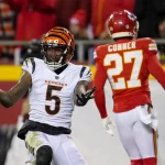 FootballR - NFL - Ein Footballspieler der Cincinnati Bengals mit der Nummer 5 gestikuliert mit offenen, nach oben gerichteten Händen. Er steht auf einem Footballfeld, und ein Spieler der Kansas City Chiefs mit der Nummer 27 ist abgewandt und geht in die entgegengesetzte Richtung. Im Hintergrund ist eine Menschenmenge auf den Tribünen zu sehen und es sind Andeutungen von Franchise-Ausschreibungsverhandlungen mit Tee Higgins zu erkennen. Diese Beschreibung wurde automatisch generiert.