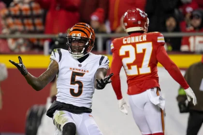 FootballR - NFL - Ein Footballspieler der Cincinnati Bengals mit der Nummer 5 gestikuliert mit offenen, nach oben gerichteten Händen. Er steht auf einem Footballfeld, und ein Spieler der Kansas City Chiefs mit der Nummer 27 ist abgewandt und geht in die entgegengesetzte Richtung. Im Hintergrund ist eine Menschenmenge auf den Tribünen zu sehen und es sind Andeutungen von Franchise-Ausschreibungsverhandlungen mit Tee Higgins zu erkennen. Diese Beschreibung wurde automatisch generiert.