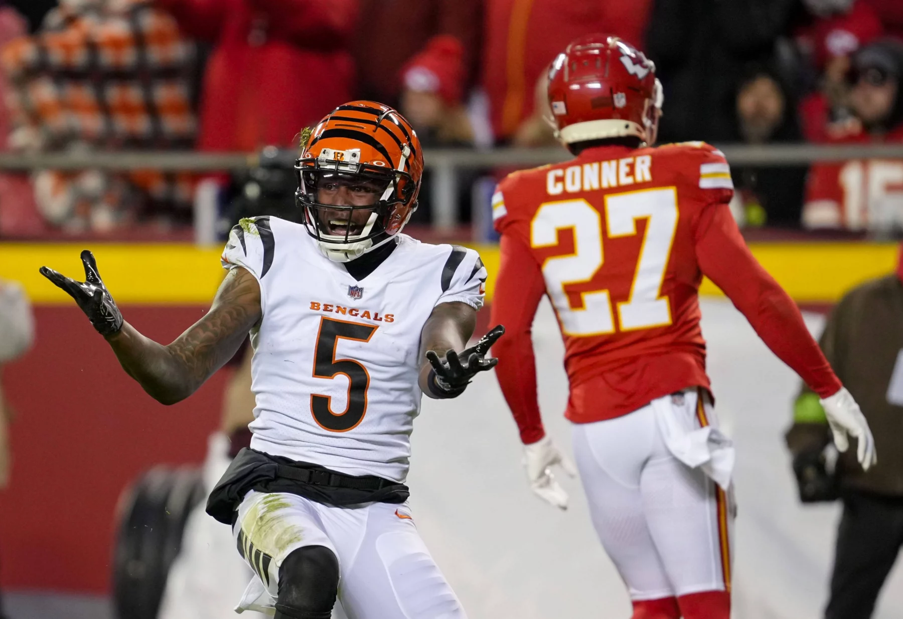 FootballR - NFL - Ein Footballspieler der Cincinnati Bengals mit der Nummer 5 gestikuliert mit offenen, nach oben gerichteten Händen. Er steht auf einem Footballfeld, und ein Spieler der Kansas City Chiefs mit der Nummer 27 ist abgewandt und geht in die entgegengesetzte Richtung. Im Hintergrund ist eine Menschenmenge auf den Tribünen zu sehen und es sind Andeutungen von Franchise-Ausschreibungsverhandlungen mit Tee Higgins zu erkennen. Diese Beschreibung wurde automatisch generiert.