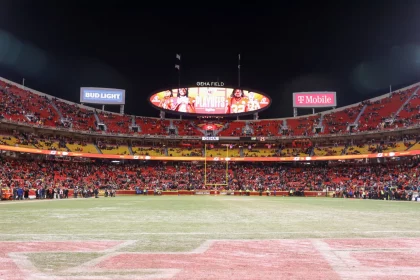 FootballR - NFL- GEHA Field - Ein Panoramablick auf ein Footballstadion mit einer großen Anzeigetafel, auf der „Playoffs“ und Bilder von Spielern angezeigt werden. Die Sitze im Stadion sind mit Fans besetzt, einige stehen, und verschiedene Werbespots wie Bud Light und T-Mobile sind zu sehen. Auf dem Spielfeld sind Schneeflecken zu sehen, und in der Ferne sind Torpfosten zu sehen, die an denkwürdige Spiele der Chiefs erinnern. Diese Beschreibung wurde automatisch generiert.