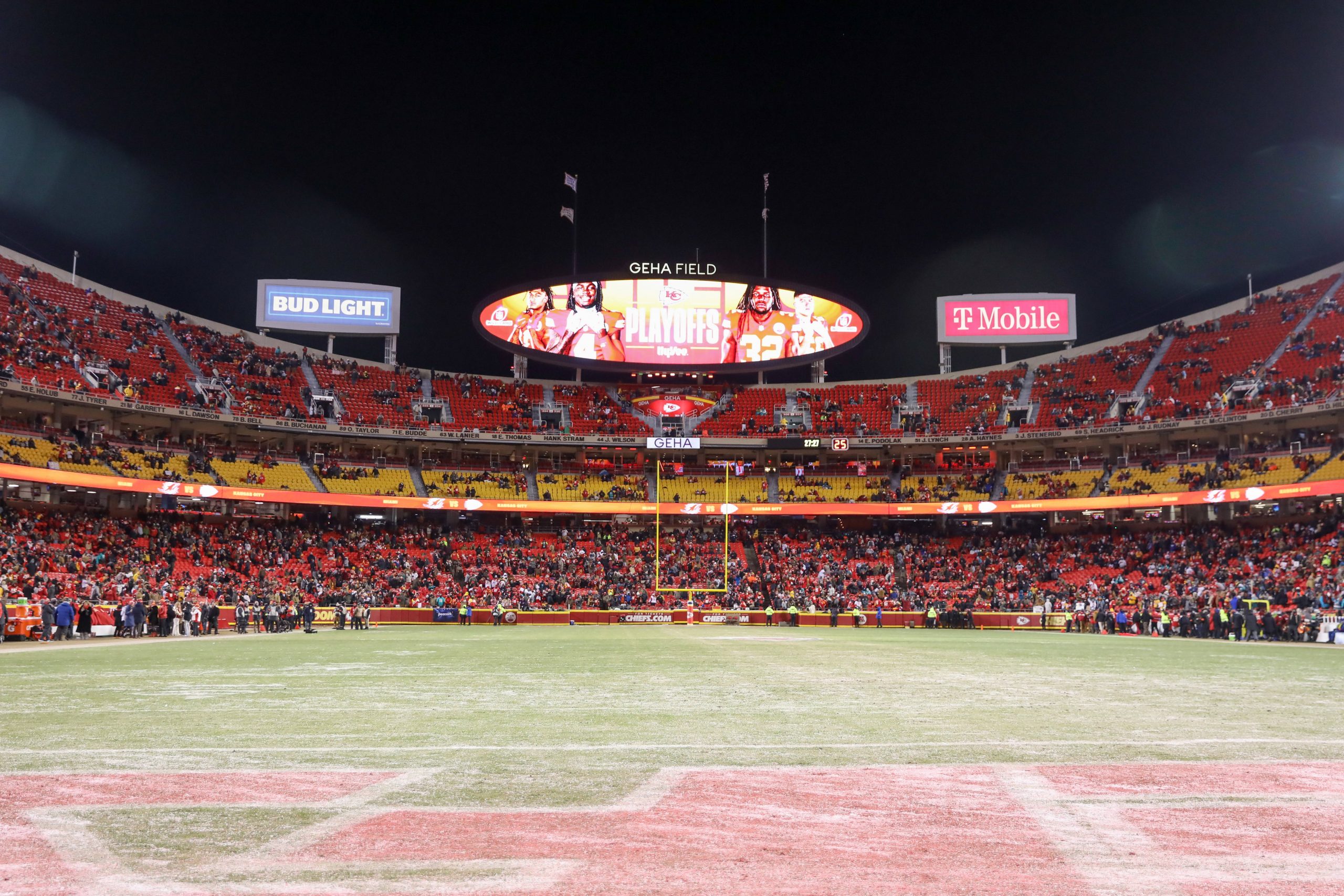 FootballR - NFL- GEHA Field - Ein Panoramablick auf ein Footballstadion mit einer großen Anzeigetafel, auf der „Playoffs“ und Bilder von Spielern angezeigt werden. Die Sitze im Stadion sind mit Fans besetzt, einige stehen, und verschiedene Werbespots wie Bud Light und T-Mobile sind zu sehen. Auf dem Spielfeld sind Schneeflecken zu sehen, und in der Ferne sind Torpfosten zu sehen, die an denkwürdige Spiele der Chiefs erinnern. Diese Beschreibung wurde automatisch generiert.