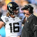 FootballR - NFL - Ein Footballspieler, Trevor Lawrence, trägt eine Uniform der Jacksonville Jaguars und einen Helm mit der Nummer 16. Er steht auf dem Feld und hört einem Trainer zu, der eine schwarze Jacke und ein Headset trägt. Die Hände des Spielers ruhen auf seinen Hüften, während sie während des Spiels einen Spielzug besprechen. Diese Beschreibung wurde automatisch generiert.