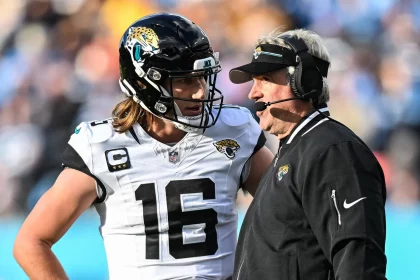 FootballR - NFL - Ein Footballspieler, Trevor Lawrence, trägt eine Uniform der Jacksonville Jaguars und einen Helm mit der Nummer 16. Er steht auf dem Feld und hört einem Trainer zu, der eine schwarze Jacke und ein Headset trägt. Die Hände des Spielers ruhen auf seinen Hüften, während sie während des Spiels einen Spielzug besprechen. Diese Beschreibung wurde automatisch generiert.
