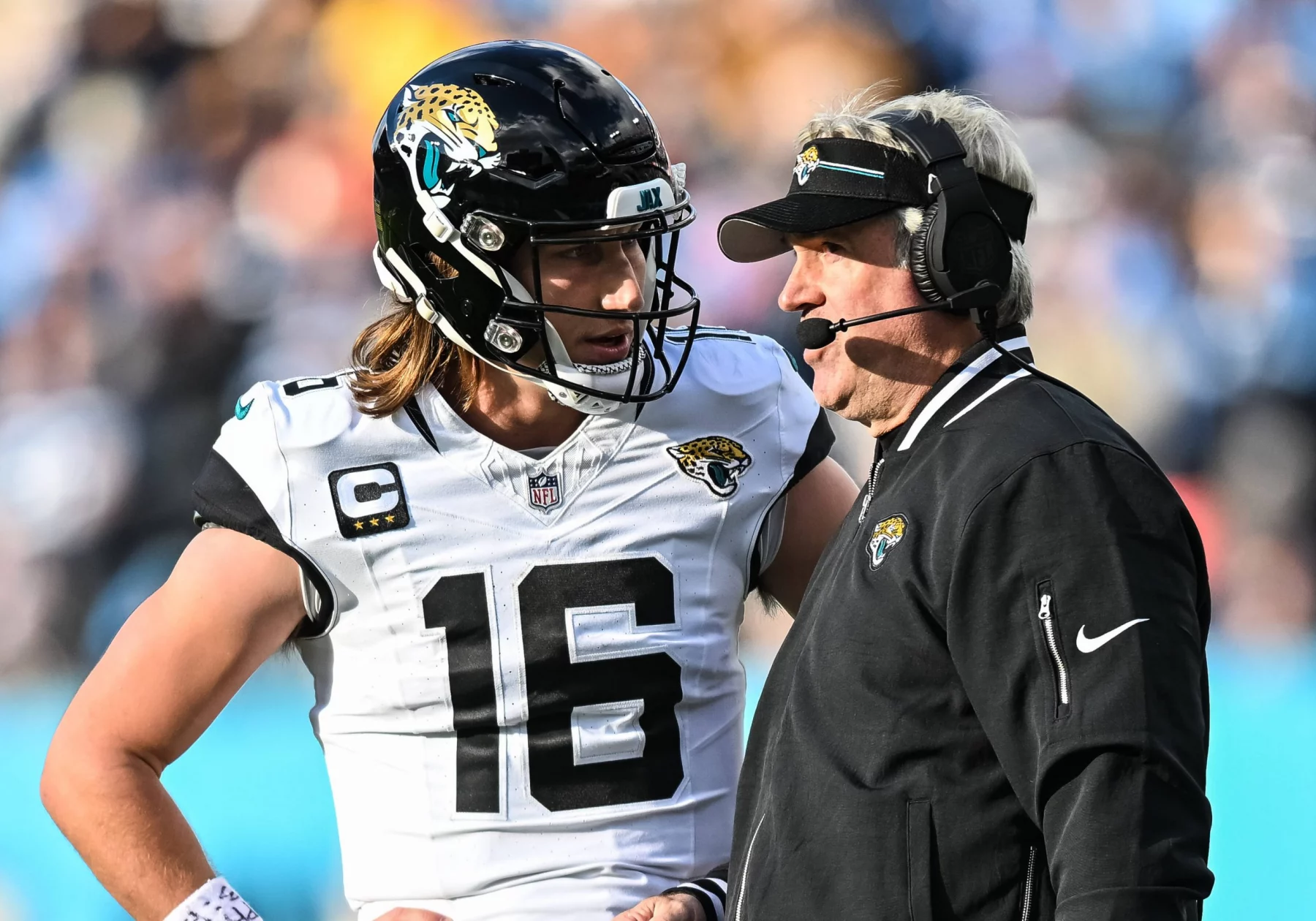 FootballR - NFL - Ein Footballspieler, Trevor Lawrence, trägt eine Uniform der Jacksonville Jaguars und einen Helm mit der Nummer 16. Er steht auf dem Feld und hört einem Trainer zu, der eine schwarze Jacke und ein Headset trägt. Die Hände des Spielers ruhen auf seinen Hüften, während sie während des Spiels einen Spielzug besprechen. Diese Beschreibung wurde automatisch generiert.