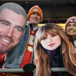 FootballR - NFL Travis Kelce - NFL International Fan of the Year - Drei Fans stehen in einem Stadion und halten große Ausschnitte von Gesichtern. Der mittlere Fan, ein Mann in einem roten Trikot und einer rot-gelben Mütze, hält den Ausschnitt des Gesichts eines bärtigen Mannes. Ihn flankieren zwei Frauen in roter Kansas City Chiefs-Ausrüstung, die Ausschnitte von Taylor Swifts Gesicht mit langen braunen Haaren halten. Im Hintergrund sind weitere Fans und die Lichter des Stadions zu sehen. Diese Beschreibung wurde automatisch generiert.