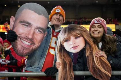 FootballR - NFL Travis Kelce - NFL International Fan of the Year - Drei Fans stehen in einem Stadion und halten große Ausschnitte von Gesichtern. Der mittlere Fan, ein Mann in einem roten Trikot und einer rot-gelben Mütze, hält den Ausschnitt des Gesichts eines bärtigen Mannes. Ihn flankieren zwei Frauen in roter Kansas City Chiefs-Ausrüstung, die Ausschnitte von Taylor Swifts Gesicht mit langen braunen Haaren halten. Im Hintergrund sind weitere Fans und die Lichter des Stadions zu sehen. Diese Beschreibung wurde automatisch generiert.