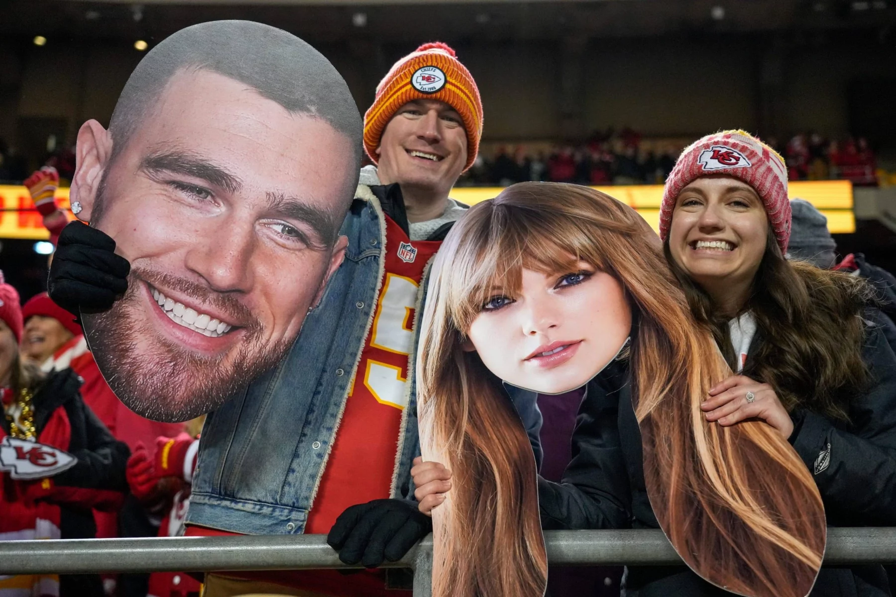 FootballR - NFL Travis Kelce - NFL International Fan of the Year - Drei Fans stehen in einem Stadion und halten große Ausschnitte von Gesichtern. Der mittlere Fan, ein Mann in einem roten Trikot und einer rot-gelben Mütze, hält den Ausschnitt des Gesichts eines bärtigen Mannes. Ihn flankieren zwei Frauen in roter Kansas City Chiefs-Ausrüstung, die Ausschnitte von Taylor Swifts Gesicht mit langen braunen Haaren halten. Im Hintergrund sind weitere Fans und die Lichter des Stadions zu sehen. Diese Beschreibung wurde automatisch generiert.
