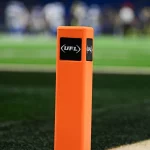 UFL Debakel - NFL - Nahaufnahme eines orangefarbenen Pylons mit dem UFL -Logo auf einem Footballfeld. Der grüne Rasen ist sichtbar, mit weißen und schwarzen Feldmarkierungen. Im Hintergrund sind verschwommene Spieler in Uniformen zu sehen. Der Fokus liegt auf dem Pylon, der die Textur und die Logos hervorhebt und eine weitere erfolgreiche Saison für Zuseher symbolisiert. Diese Beschreibung wurde automatisch generiert.