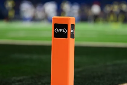 UFL Debakel - NFL - Nahaufnahme eines orangefarbenen Pylons mit dem UFL -Logo auf einem Footballfeld. Der grüne Rasen ist sichtbar, mit weißen und schwarzen Feldmarkierungen. Im Hintergrund sind verschwommene Spieler in Uniformen zu sehen. Der Fokus liegt auf dem Pylon, der die Textur und die Logos hervorhebt und eine weitere erfolgreiche Saison für Zuseher symbolisiert. Diese Beschreibung wurde automatisch generiert.