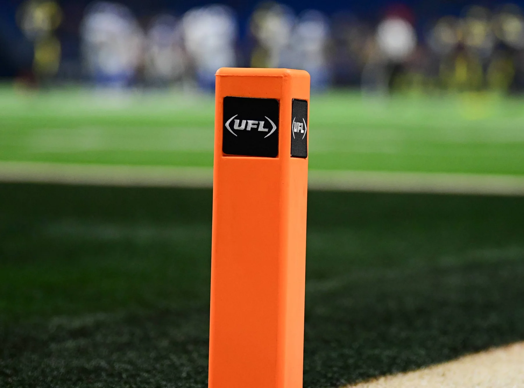 UFL Debakel - NFL - Nahaufnahme eines orangefarbenen Pylons mit dem UFL -Logo auf einem Footballfeld. Der grüne Rasen ist sichtbar, mit weißen und schwarzen Feldmarkierungen. Im Hintergrund sind verschwommene Spieler in Uniformen zu sehen. Der Fokus liegt auf dem Pylon, der die Textur und die Logos hervorhebt und eine weitere erfolgreiche Saison für Zuseher symbolisiert. Diese Beschreibung wurde automatisch generiert.
