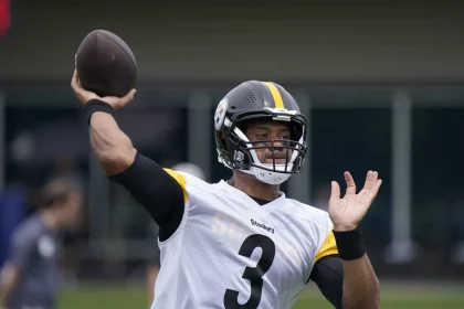 FootballR - 4 NFL-Stars - Ein Footballspieler mit einem schwarzen Helm mit einem gelben Streifen und der Nummer 3, der an Russell Wilson Stil erinnert, wirft einen Football. Er trägt ein weißes Trikot mit schwarzen und gelben Ärmeln. Im Hintergrund sind verschwommene Figuren und ein grünes Feld zu sehen, was an eine Trainingseinheit der neugeborenen Steelers erinnert. Diese Beschreibung wurde automatisch generiert.