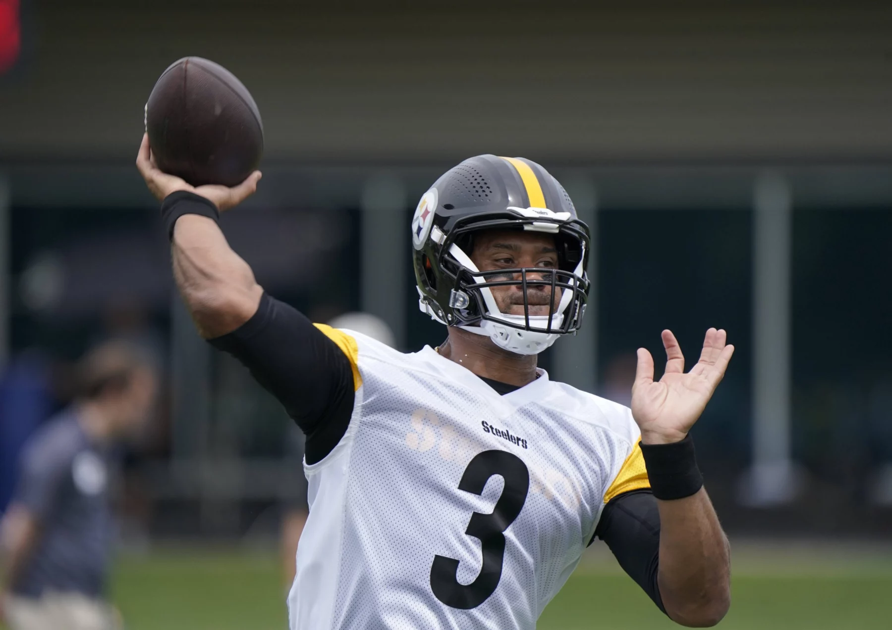 FootballR - 4 NFL-Stars - Ein Footballspieler mit einem schwarzen Helm mit einem gelben Streifen und der Nummer 3, der an Russell Wilson Stil erinnert, wirft einen Football. Er trägt ein weißes Trikot mit schwarzen und gelben Ärmeln. Im Hintergrund sind verschwommene Figuren und ein grünes Feld zu sehen, was an eine Trainingseinheit der neugeborenen Steelers erinnert. Diese Beschreibung wurde automatisch generiert.