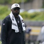 FootballR - NFL - Mike Tomlin - Ein Mann mit weißer Baseballkappe, schwarzer Sonnenbrille und schwarzer Jacke steht draußen. Um seinen Hals hängt ein weißes Handtuch, auf dem die Logos der Pittsburgh Steelers zu sehen sind. Außerdem trägt er eine silberne Halskette und ein Schlüsselband. Der Hintergrund ist verschwommen und man sieht in der Ferne Grün und Menschen. Diese Beschreibung wurde automatisch generiert.