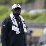 FootballR - NFL - Mike Tomlin - Ein Mann mit weißer Baseballkappe, schwarzer Sonnenbrille und schwarzer Jacke steht draußen. Um seinen Hals hängt ein weißes Handtuch, auf dem die Logos der Pittsburgh Steelers zu sehen sind. Außerdem trägt er eine silberne Halskette und ein Schlüsselband. Der Hintergrund ist verschwommen und man sieht in der Ferne Grün und Menschen. Diese Beschreibung wurde automatisch generiert.