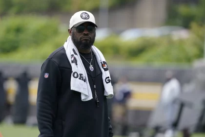 FootballR - NFL - Mike Tomlin - Ein Mann mit weißer Baseballkappe, schwarzer Sonnenbrille und schwarzer Jacke steht draußen. Um seinen Hals hängt ein weißes Handtuch, auf dem die Logos der Pittsburgh Steelers zu sehen sind. Außerdem trägt er eine silberne Halskette und ein Schlüsselband. Der Hintergrund ist verschwommen und man sieht in der Ferne Grün und Menschen. Diese Beschreibung wurde automatisch generiert.
