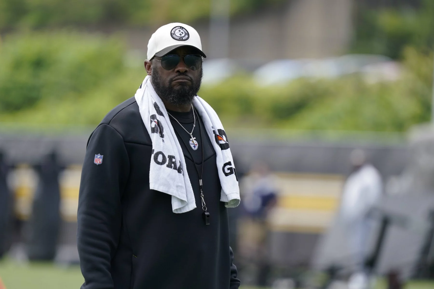 FootballR - NFL - Mike Tomlin - Ein Mann mit weißer Baseballkappe, schwarzer Sonnenbrille und schwarzer Jacke steht draußen. Um seinen Hals hängt ein weißes Handtuch, auf dem die Logos der Pittsburgh Steelers zu sehen sind. Außerdem trägt er eine silberne Halskette und ein Schlüsselband. Der Hintergrund ist verschwommen und man sieht in der Ferne Grün und Menschen. Diese Beschreibung wurde automatisch generiert.