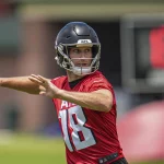 FootballR - NFL - Kirk Cousins - Ein Footballspieler in einem roten Trikot mit der Nummer 18 und einem schwarzen Helm bereitet sich während eines Trainings der Atlanta Falcons darauf vor, einen Football zu werfen. Der unscharfe Hintergrund zeigt eine digitale Spieluhr und Feldausrüstung, was auf eine Trainingsumgebung im Freien hindeutet. Diese Beschreibung wurde automatisch generiert.