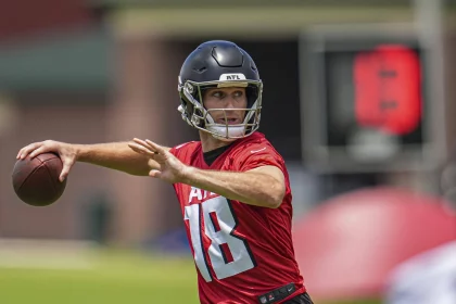 FootballR - NFL - Kirk Cousins - Ein Footballspieler in einem roten Trikot mit der Nummer 18 und einem schwarzen Helm bereitet sich während eines Trainings der Atlanta Falcons darauf vor, einen Football zu werfen. Der unscharfe Hintergrund zeigt eine digitale Spieluhr und Feldausrüstung, was auf eine Trainingsumgebung im Freien hindeutet. Diese Beschreibung wurde automatisch generiert.