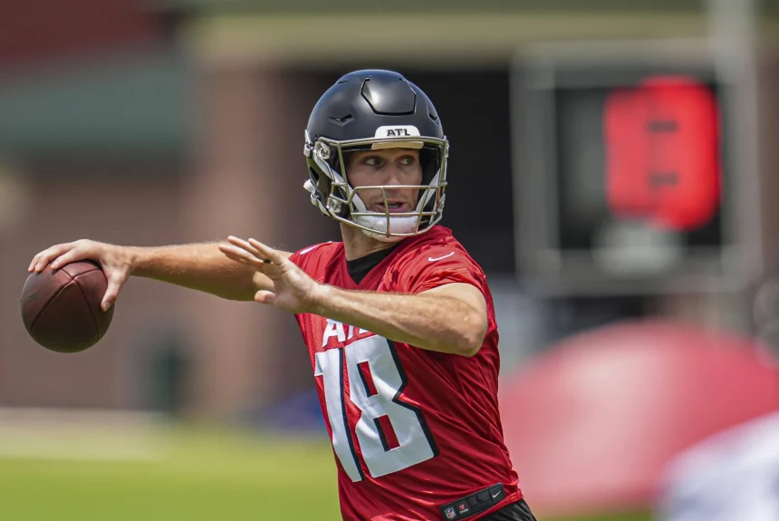 FootballR - NFL - Kirk Cousins - Ein Footballspieler in einem roten Trikot mit der Nummer 18 und einem schwarzen Helm bereitet sich während eines Trainings der Atlanta Falcons darauf vor, einen Football zu werfen. Der unscharfe Hintergrund zeigt eine digitale Spieluhr und Feldausrüstung, was auf eine Trainingsumgebung im Freien hindeutet. Diese Beschreibung wurde automatisch generiert.