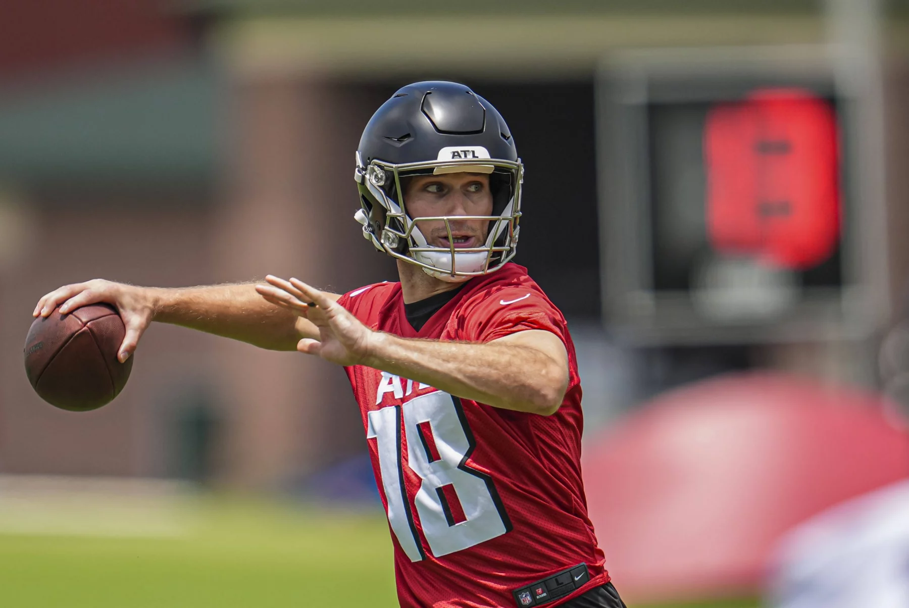 FootballR - NFL - Kirk Cousins - Ein Footballspieler in einem roten Trikot mit der Nummer 18 und einem schwarzen Helm bereitet sich während eines Trainings der Atlanta Falcons darauf vor, einen Football zu werfen. Der unscharfe Hintergrund zeigt eine digitale Spieluhr und Feldausrüstung, was auf eine Trainingsumgebung im Freien hindeutet. Diese Beschreibung wurde automatisch generiert.