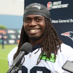 FootballR - NFL - Ein Footballspieler, Rhamondre Stevenson, trägt ein Trikot der New England Patriots und ein schwarzes Stirnband mit dem Logo des Teams und lächelt, während er bei einer Pressekonferenz vor einem Mikrofon steht. Im Hintergrund sind verschiedene Sponsorenlogos zu sehen, darunter Waste Innovations und Mass General Brigham. Diese Beschreibung wurde automatisch generiert.