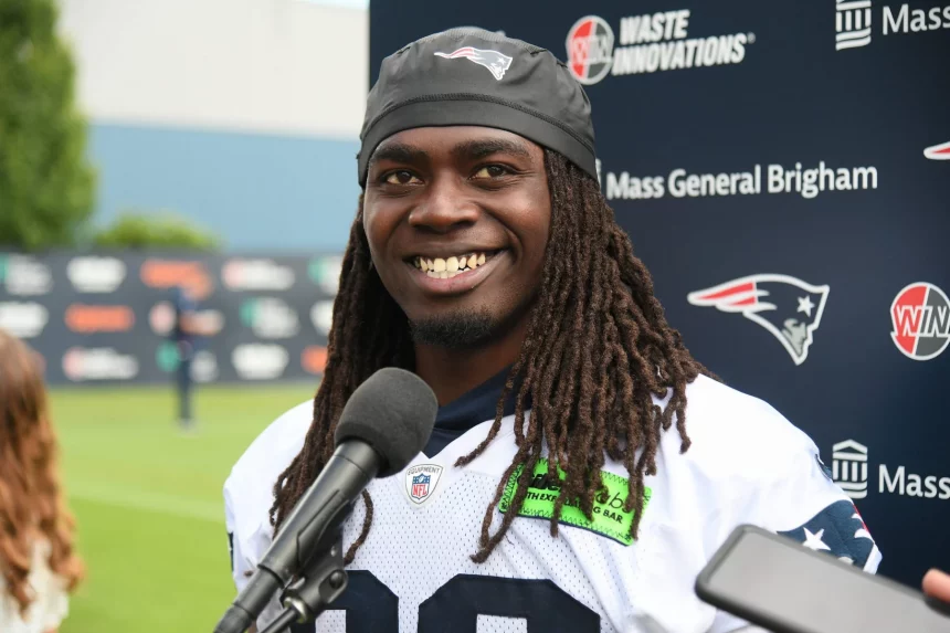 FootballR - NFL - Ein Footballspieler, Rhamondre Stevenson, trägt ein Trikot der New England Patriots und ein schwarzes Stirnband mit dem Logo des Teams und lächelt, während er bei einer Pressekonferenz vor einem Mikrofon steht. Im Hintergrund sind verschiedene Sponsorenlogos zu sehen, darunter Waste Innovations und Mass General Brigham. Diese Beschreibung wurde automatisch generiert.