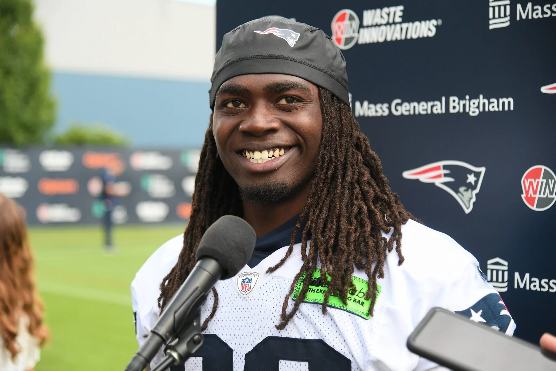 FootballR - NFL - Ein Footballspieler, Rhamondre Stevenson, trägt ein Trikot der New England Patriots und ein schwarzes Stirnband mit dem Logo des Teams und lächelt, während er bei einer Pressekonferenz vor einem Mikrofon steht. Im Hintergrund sind verschiedene Sponsorenlogos zu sehen, darunter Waste Innovations und Mass General Brigham. Diese Beschreibung wurde automatisch generiert.