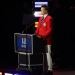 FootballR - NFL - Ein Mann steht lächelnd an einem Podium und trägt einen leuchtend roten Blazer und weiße Hosen. Auf dem Podium sind die Nummer 12 und das Wort „BRADY“ zu sehen. Im schwach beleuchteten Hintergrund mit bunten vertikalen Lichtern ist klar, dass er die Patriots-Legende Tom Brady geehrt wird. Diese Beschreibung wurde automatisch generiert.