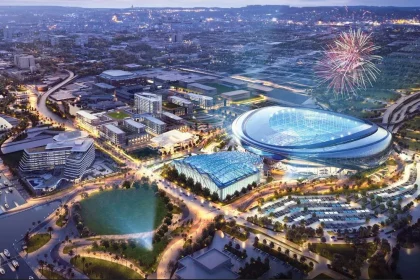 FootballR - NFL - Luftaufnahme eines modernen Stadtgebiets in der Abenddämmerung, darunter das neue NFL-Stadion der Jacksonville Jaguars mit einem durchscheinenden Dach. Die umliegenden Gebäude und Straßen sind gut beleuchtet. Am Himmel über dem Stadion explodieren Feuerwerke. In der Umgebung befinden sich ein Gewässer, Grünflächen und ein Yachthafen mit Booten. Diese Beschreibung wurde automatisch generiert.