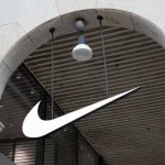 FootballR - NFL - Ein großes, weißes Nike Swoosh Logo hängt unter einem Steintorbogen vor einem Geschäft. Der Torbogen hat eine beige Fassade mit sichtbaren Steinstrukturen. Über dem Logo, das mit ihrer NFL-Partnerschaft in Verbindung steht, ist an der Decke, die mit horizontalen Holzlamellen versehen ist, eine einzelne weiße Überwachungskamera mit eingebauter Leuchte angebracht. Diese Beschreibung wurde automatisch generiert.