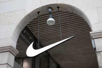 FootballR - NFL - Ein großes, weißes Nike Swoosh Logo hängt unter einem Steintorbogen vor einem Geschäft. Der Torbogen hat eine beige Fassade mit sichtbaren Steinstrukturen. Über dem Logo, das mit ihrer NFL-Partnerschaft in Verbindung steht, ist an der Decke, die mit horizontalen Holzlamellen versehen ist, eine einzelne weiße Überwachungskamera mit eingebauter Leuchte angebracht. Diese Beschreibung wurde automatisch generiert.